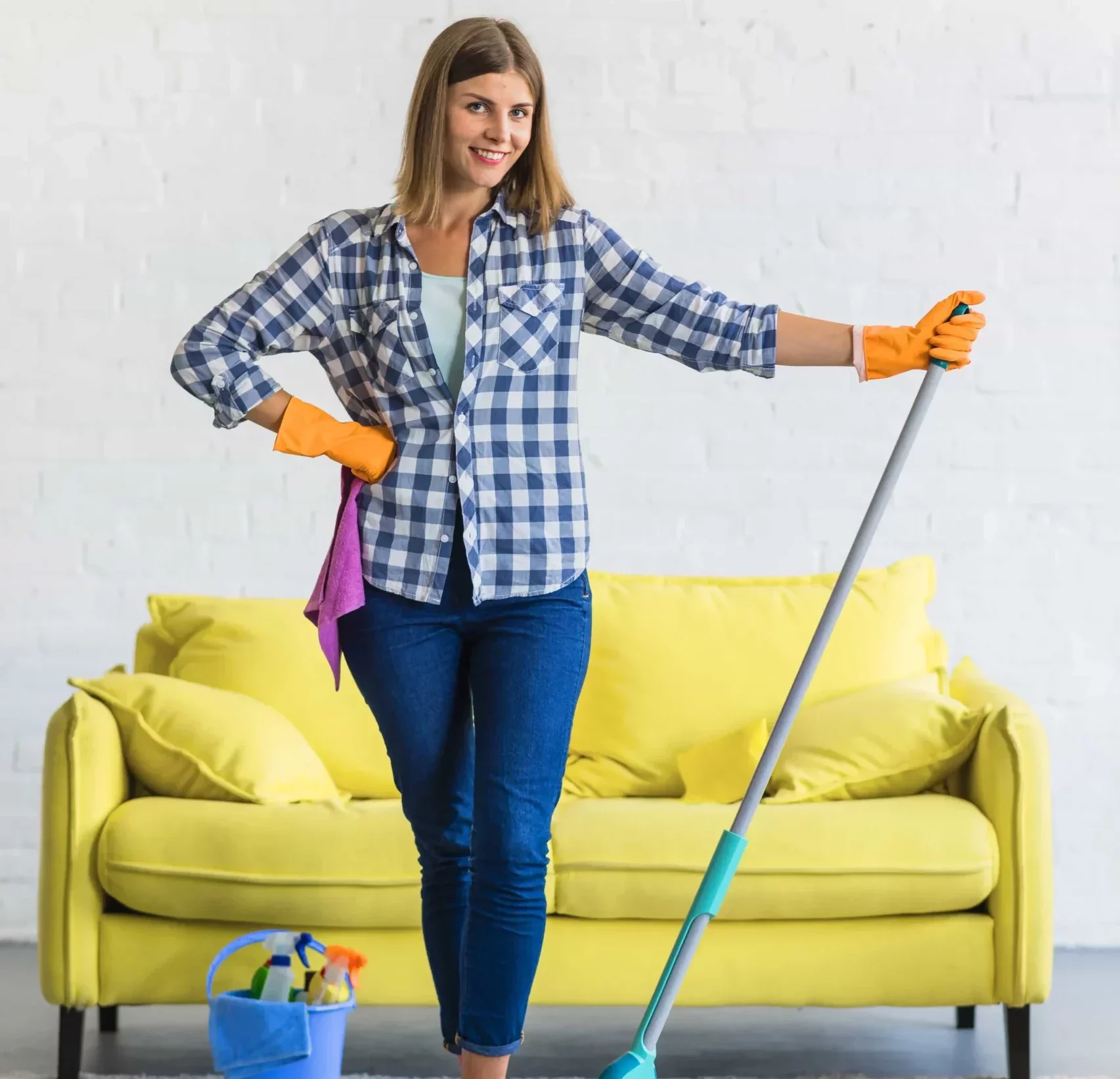 smiling young woman holding a mop while standing in front of a sofa next to a bucket of cleaning products