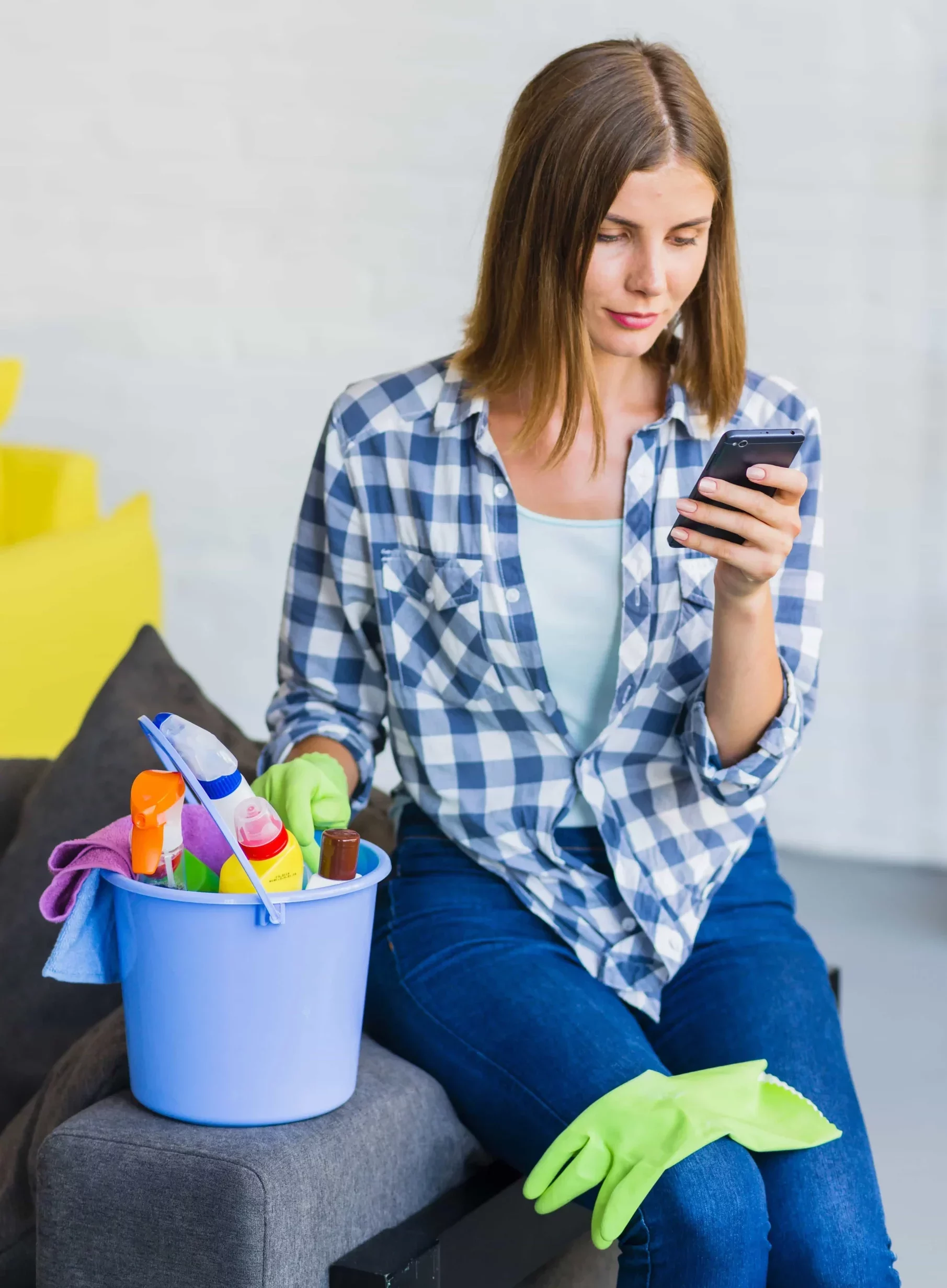 woman sitting on the couch holding a bucket full of cleaning products while looking at her phone screen