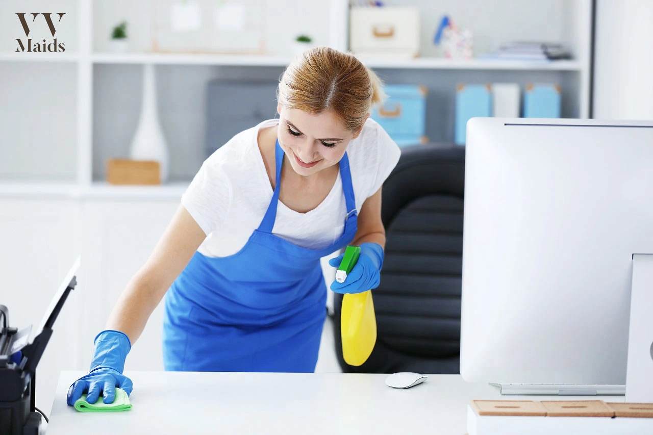 A professional cleaner from VV Maids Cleaning Services in NY wipes down a desk with cleaning supplies in a tidy office