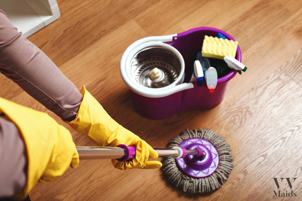 A person in yellow gloves uses a mop next to a purple cleaning bucket, representing VV Maids Cleaning Services in NY.