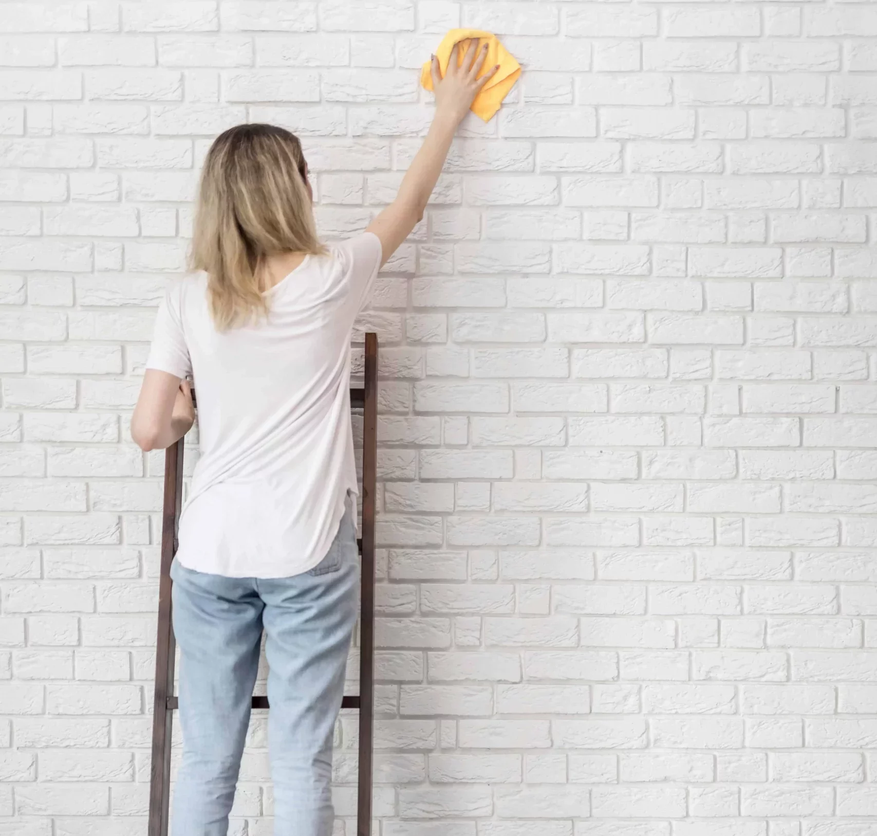 woman standing on a ladder to clean the wall with a yellow cleaning cloth
