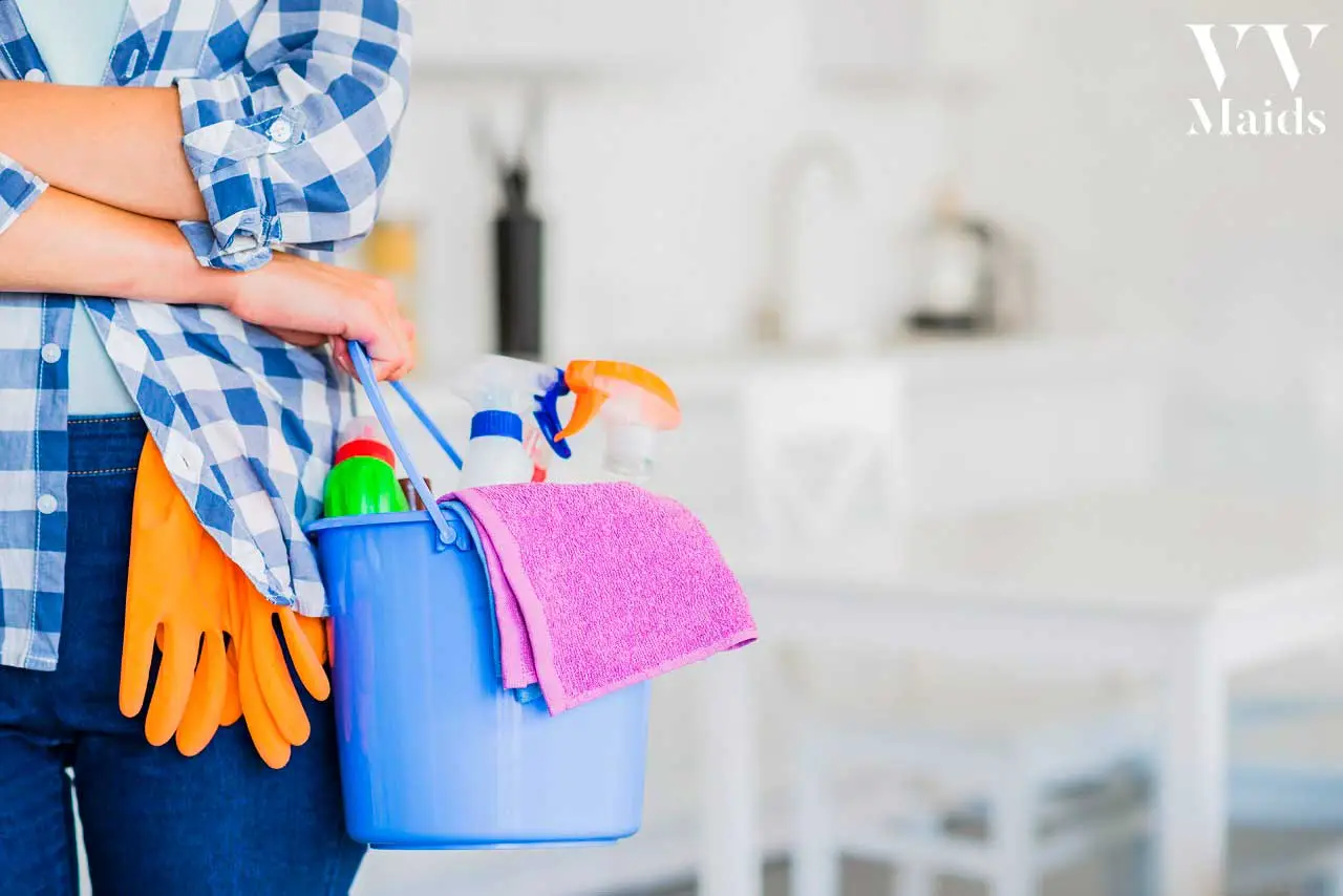 woman in a checkered pattern shirt holding a blue bucket full of cleaning products with orange cleaning gloves